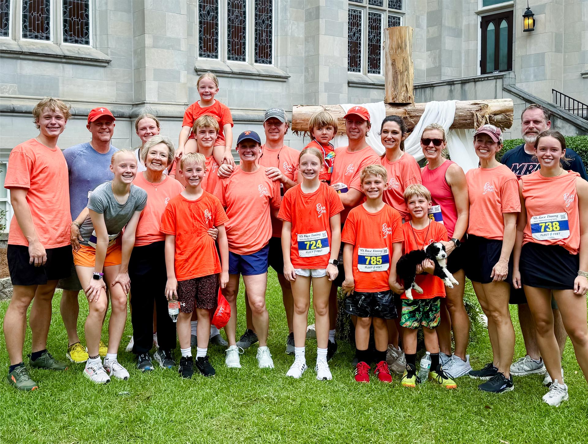 Photo of large family with grandmother, adult children, and grandchildren in front of cross at First Baptist Jackson. One child is holding a puppy.